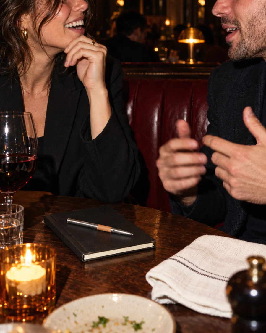 Two people talking across a restaurant table at night, the pen resting on a closed notebook